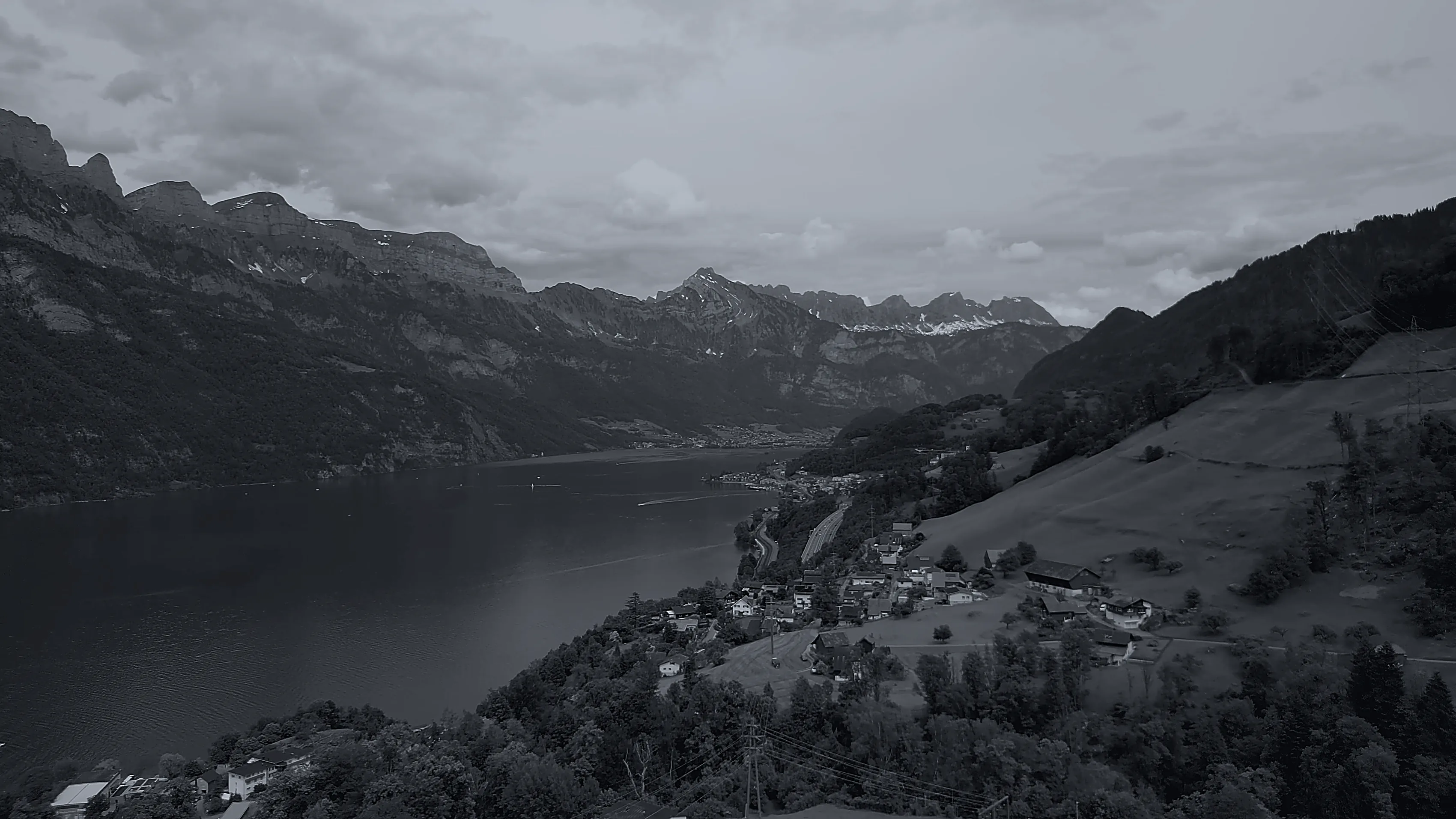 Grau getönter Blick über den Walensee: ein Dorf an einem grünen Hang über dem See, Felswände und verschneite Gipfel unter bewölktem Himmel.